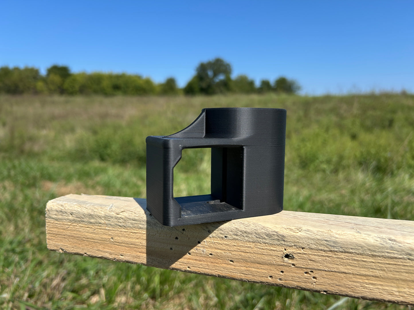 Black T-Dapter mounting bracket resting on a horizontal wooden 2×2 board outdoors, shown from the side with an open rectangular slot for mounting wood 2x2s to a metal T-post.