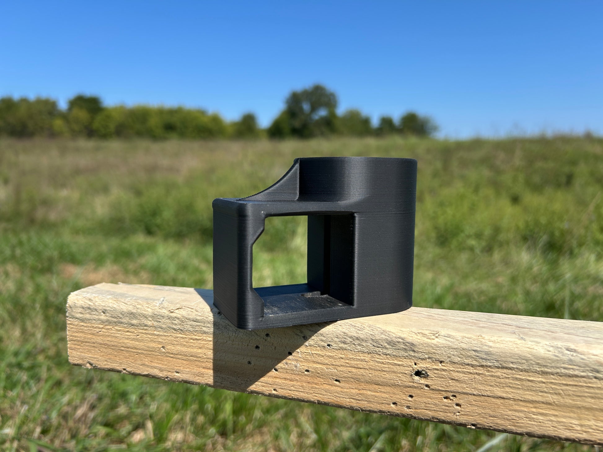 Black T-Dapter mounting bracket resting on a horizontal wooden 2×2 board outdoors, shown from the side with an open rectangular slot for mounting wood 2x2s to a metal T-post.