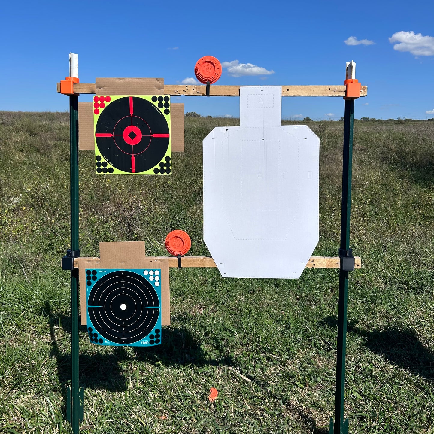 Outdoor shooting target stand constructed with vertical metal T-posts and horizontal wooden 2×2 boards, holding paper targets and orange clay targets in an open grassy field under a clear blue sky.