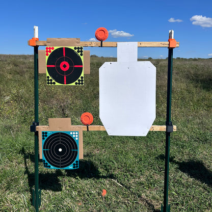Outdoor shooting target stand constructed with vertical metal T-posts and horizontal wooden 2×2 boards, holding paper targets and orange clay targets in an open grassy field under a clear blue sky.