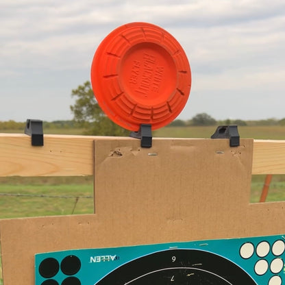 Orange clay shooting target mounted on a horizontal wooden 2×2 board above a cardboard backer, held in place by black plastic clips outdoors.
