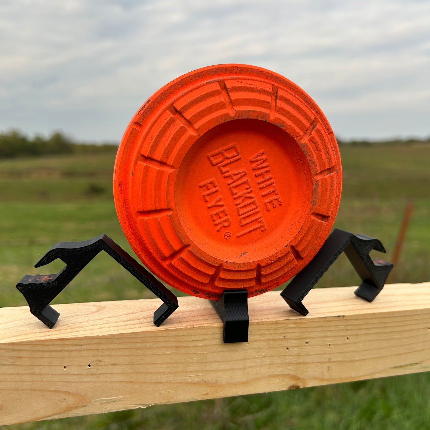 Orange clay shooting target supported on a horizontal wooden 2×2 board using angled black plastic clips outdoors in a grassy field.