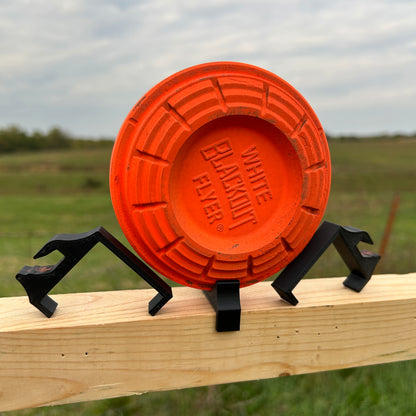 Orange clay shooting target supported on a horizontal wooden 2×2 board using angled black plastic clips outdoors in a grassy field.