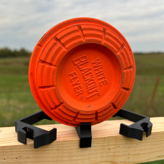 Orange clay shooting target positioned on a horizontal wooden 2×2 board with multiple black plastic clips displayed nearby outdoors.