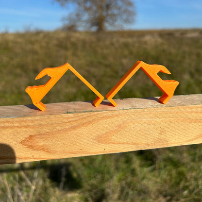 two orange plastic clips displayed on a horizontal wooden 2×2 board outdoors, shown from the side.