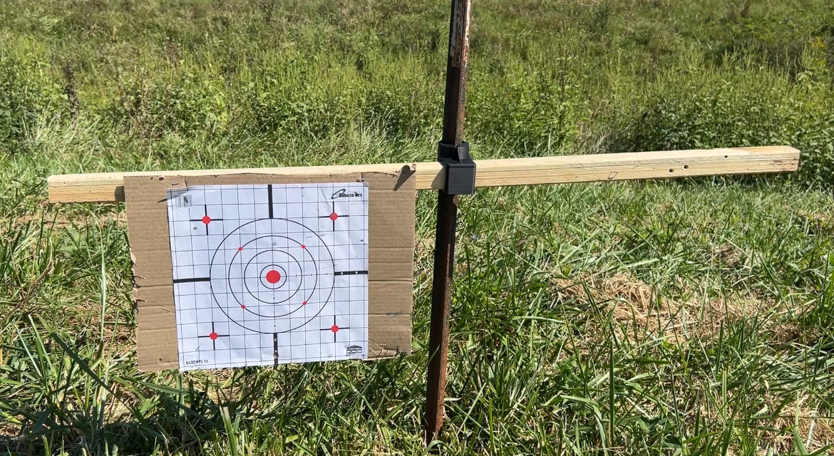 Paper shooting target attached to a horizontal wooden 2×2 board mounted to a vertical metal T-post using a black T-Dapter bracket, shown outdoors in a grassy field.