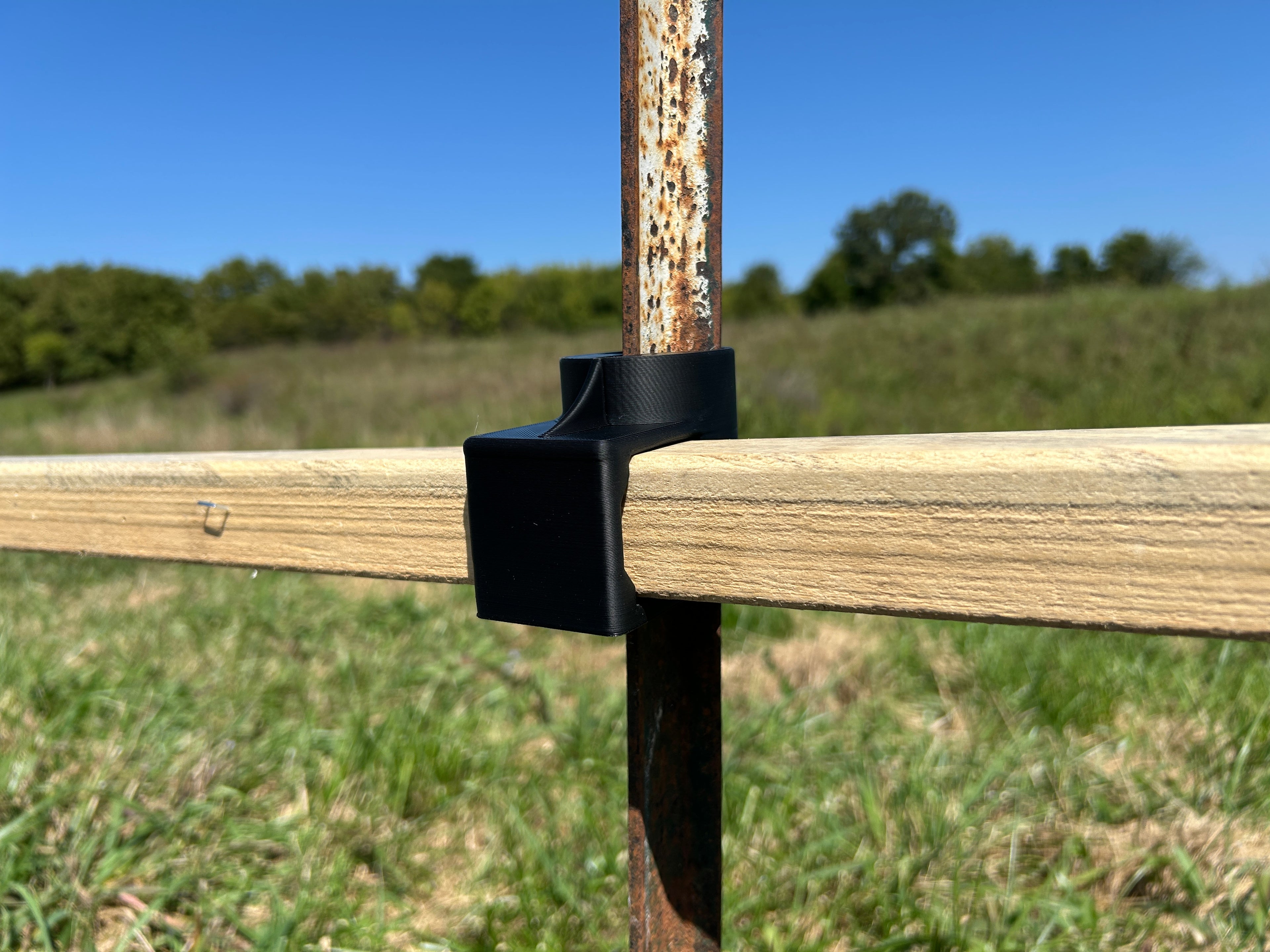 Black T-Dapter mounting bracket securing a horizontal wooden 2×2 board to a vertical metal T-post outdoors in a grassy field.