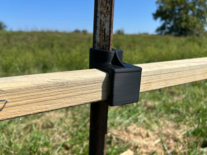 Black T-Dapter mounting bracket securing a horizontal wooden 2×2 board to a vertical metal T-post outdoors in a grassy field.