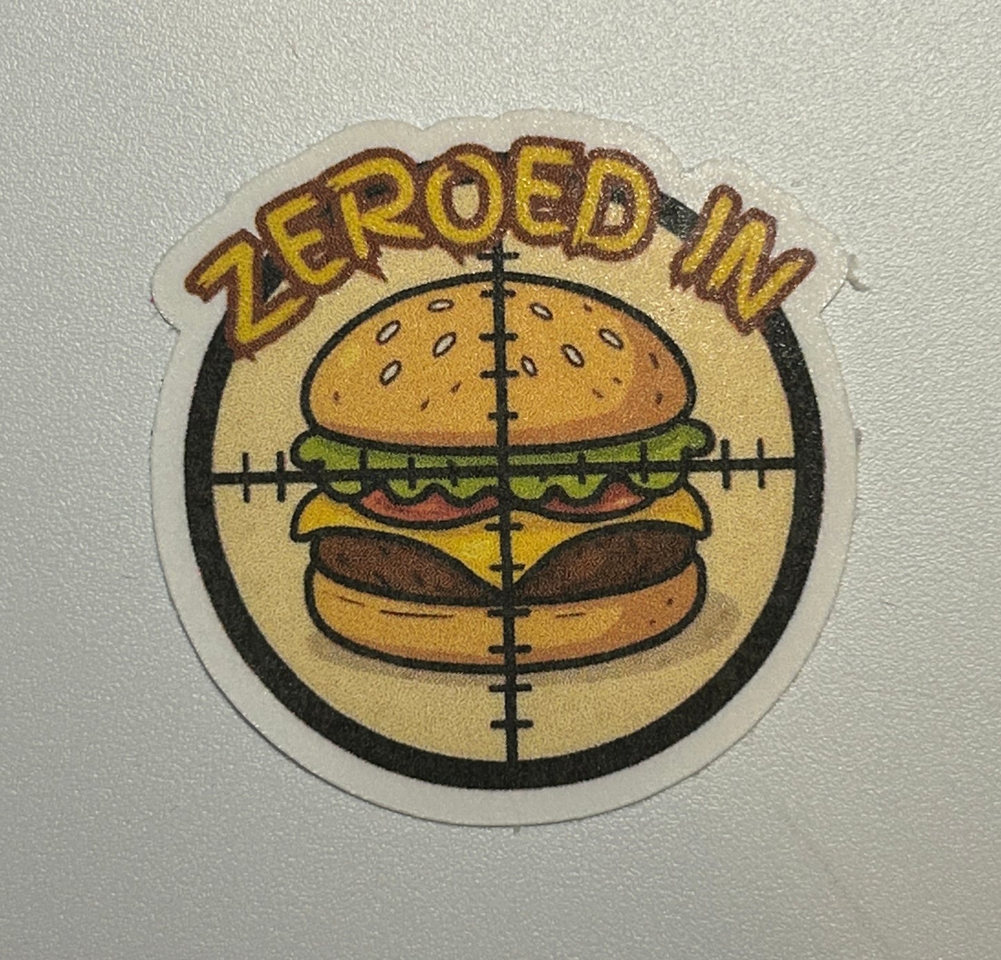 Round sticker showing an illustrated hamburger centered within a crosshair-style graphic and surrounding text.