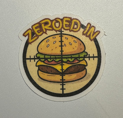 Round sticker showing an illustrated hamburger centered within a crosshair-style graphic and surrounding text.